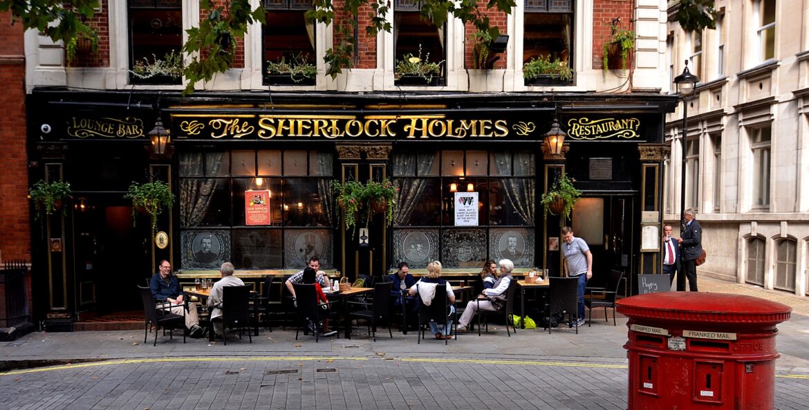 Exterior of The Sherlock Holmes pub with golden signage, black facade, and outdoor dining along a brick street. Plants hang from above.