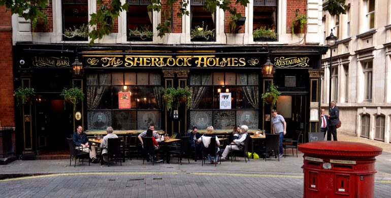 Exterior of The Sherlock Holmes pub with golden signage, black facade, and outdoor dining along a brick street. Plants hang from above.