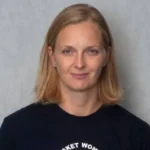 Head-and-shoulders portrait of a woman with shoulder-length blonde hair and a neutral expression, wearing a black crew-neck shirt against a gray wall.
