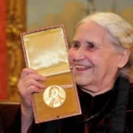 Senior woman with white hair holding an open box containing a gold medal, smiling at an awards event.