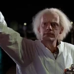 An elderly man with frizzy white hair wearing a white lab coat raises one arm in a dark night scene by the water, looking forward with a serious expression.