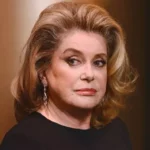 Portrait of a mature woman with styled blonde hair, wearing dangling earrings and a black top, looking to the left.