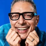 Smiling man with short gray hair and black-framed glasses against a blue background.