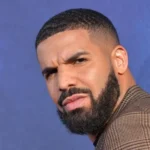 Man with a short fade and full beard, wearing a brown checkered jacket, squinting at the camera against a blue background.