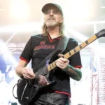 Male guitarist on a concert stage, wearing a cap and sunglasses, playing a black electric guitar.