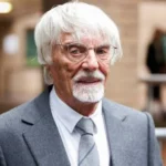 Bernie Ecclestone - NOTE.radio - Polskie radio w UK | Polskie radio w Londynie Older man with white hair and beard wearing a gray suit and glasses, posing in a modern indoor setting.