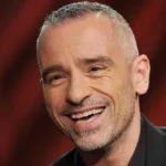 Eros Ramazzotti - NOTE.radio - Polskie radio w UK | Polskie radio w Londynie Man smiling warmly, short gray hair, wearing a dark shirt against a blurred red background.
