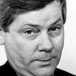 Close-up black-and-white portrait of a man with short dark hair and a neutral, slightly pensive expression, facing slightly right.