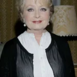 Smiling older woman with short blond hair in a white blouse and black cardigan, posing indoors against vintage wallpaper and a lamp in the background.