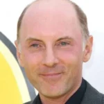 Close-up of a smiling man with a receding hairline wearing a dark blazer against a light background.