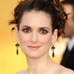 Close-up portrait of a woman with a dark updo, wearing black statement earrings, against a yellow backdrop.