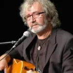 Older man with gray hair and beard, wearing glasses, playing an acoustic guitar on stage while singing into a microphone.