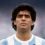 Close-up of a man with dark wavy hair wearing a light blue and white striped soccer jersey, outdoors with a serious expression.