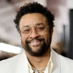 Portrait of a smiling man with curly black hair and a beard, wearing gold-rimmed glasses and a cream blazer indoors.