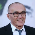 Danny Boyle - NOTE.radio - Polskie radio w UK | Polskie radio w Londynie Smiling older man wearing a black suit, white shirt, and gray tie, with rectangular glasses at a formal event.
