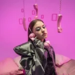 Bambi - NOTE.radio - Polskie radio w UK | Polskie radio w Londynie Young woman wearing pink headphones and a metallic jacket sits on a bed under pink lighting, with old-fashioned phone receivers hanging from the ceiling.”