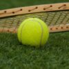 Bright yellow tennis ball resting on grass with a tennis racket in the background, on a sunny court surface.