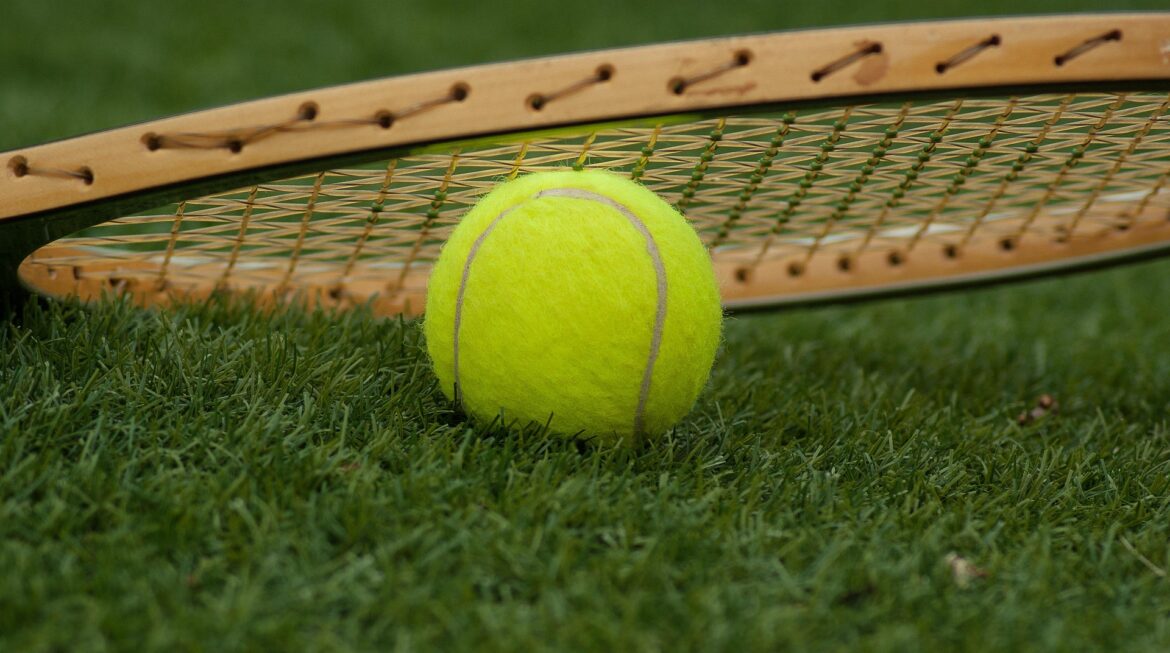 Bright yellow tennis ball resting on grass with a tennis racket in the background, on a sunny court surface.