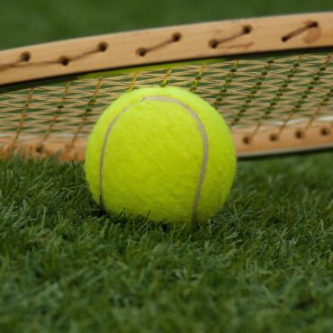 Bright yellow tennis ball resting on grass with a tennis racket in the background, on a sunny court surface.