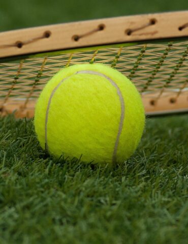 Bright yellow tennis ball resting on grass with a tennis racket in the background, on a sunny court surface.