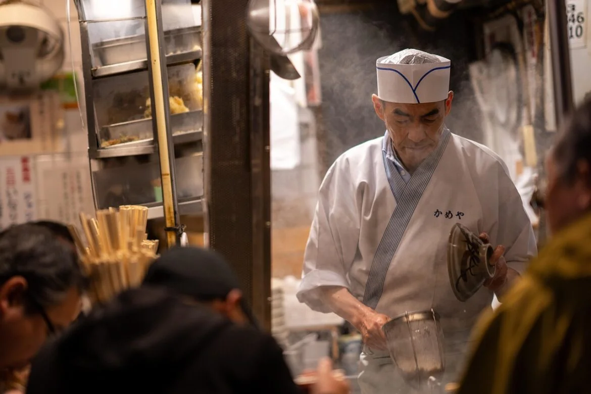 Chef in white traditional jacket and hat works at a steam-filled street food stall, holding a pot and lid while cooking behind a counter.