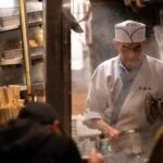 Chef in white traditional jacket and hat works at a steam-filled street food stall, holding a pot and lid while cooking behind a counter.