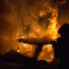Silhouetted firefighter in protective gear standing beside a large blaze with orange flames and thick smoke at night
