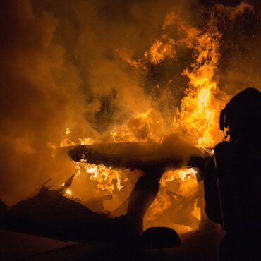 Silhouetted firefighter in protective gear standing beside a large blaze with orange flames and thick smoke at night