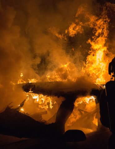 Silhouetted firefighter in protective gear standing beside a large blaze with orange flames and thick smoke at night