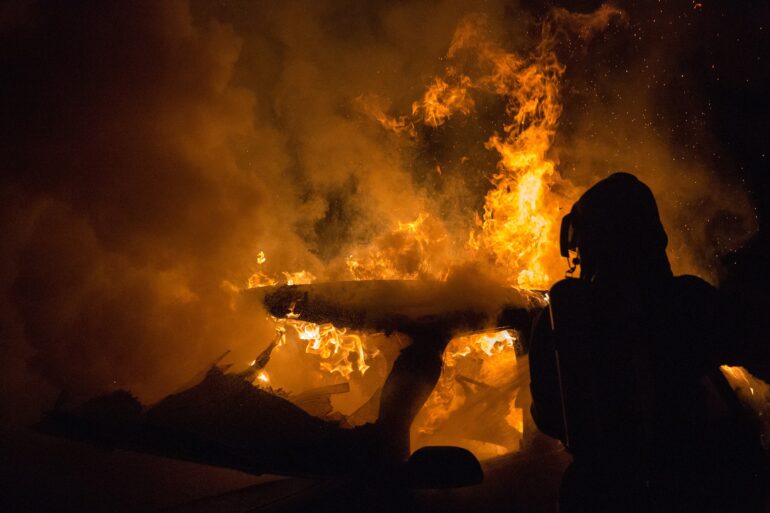 Silhouetted firefighter in protective gear standing beside a large blaze with orange flames and thick smoke at night