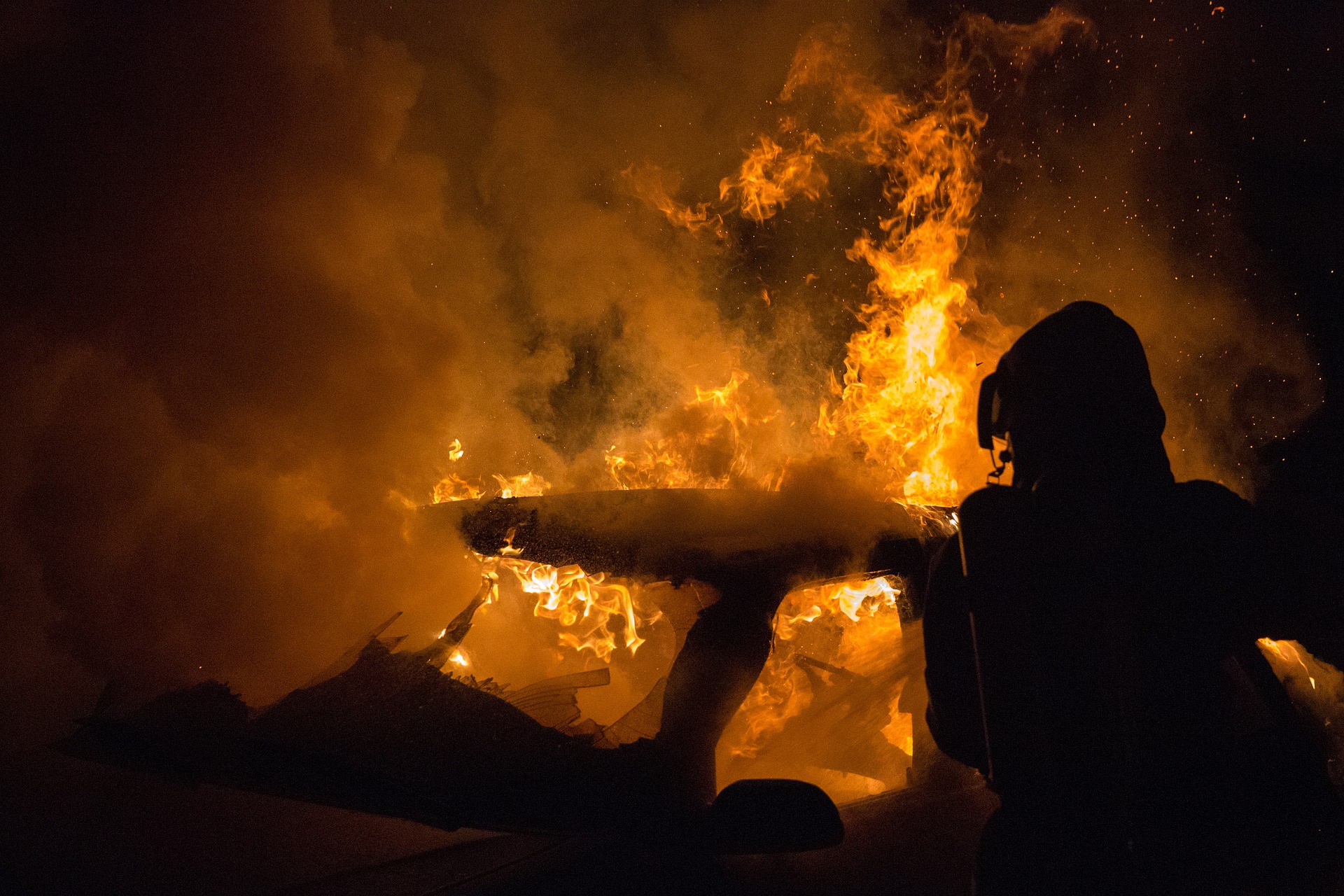 Silhouetted firefighter in protective gear standing beside a large blaze with orange flames and thick smoke at night