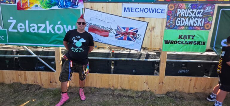 Man with pink mohawk, sunglasses, black T-shirt and pink boots posing in front of a wooden barrier with colorful banners at an outdoor festival festival scene.