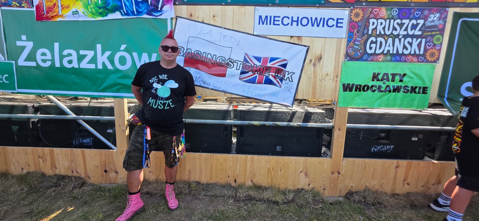 Man with pink mohawk, sunglasses, black T-shirt and pink boots posing in front of a wooden barrier with colorful banners at an outdoor festival festival scene.