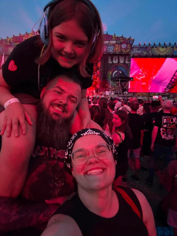 Two friends take a selfie at a large outdoor concert, smiles facing the camera with a colorful stage and cheering crowd behind them.