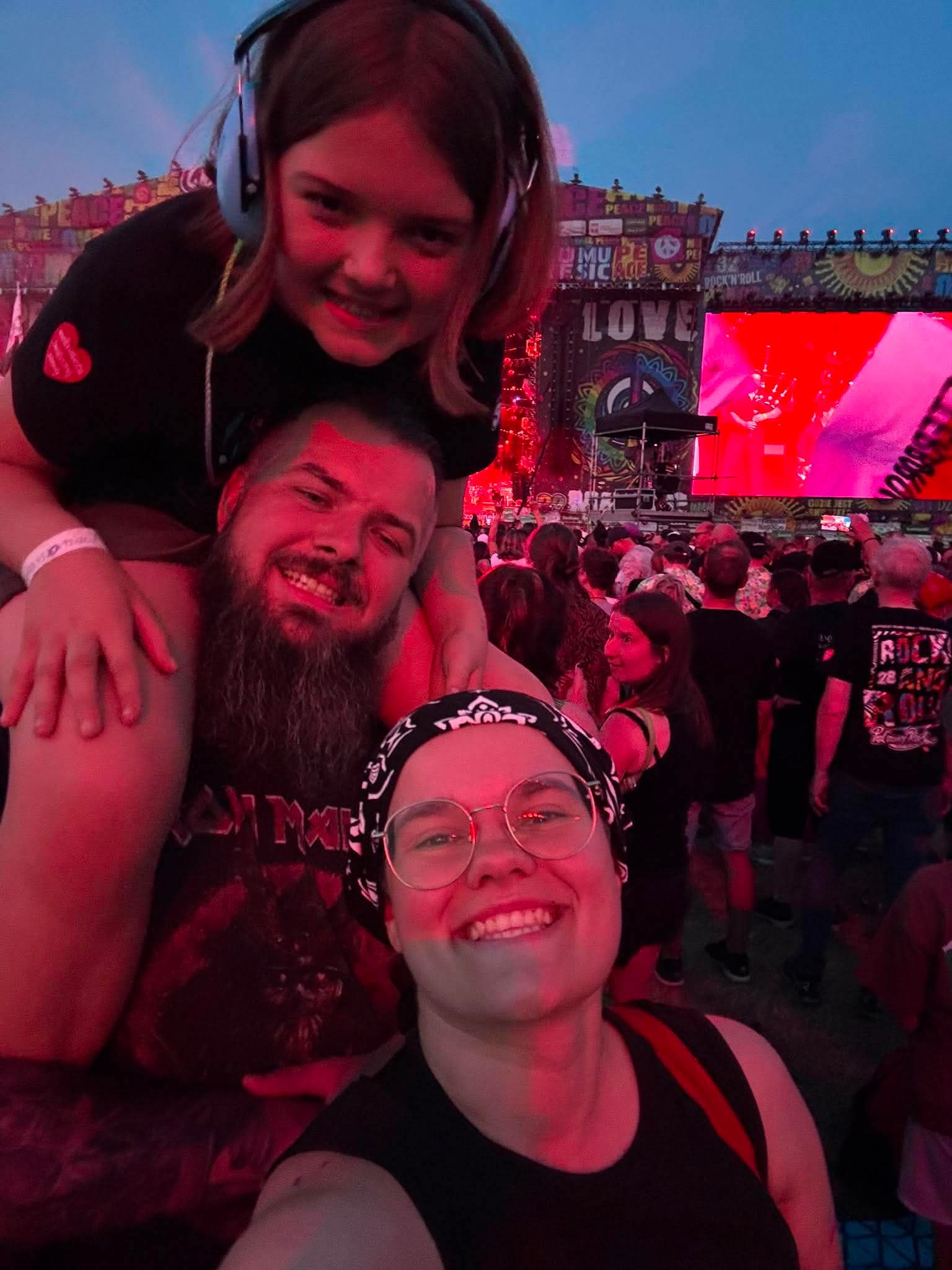 Two friends take a selfie at a large outdoor concert, smiles facing the camera with a colorful stage and cheering crowd behind them.