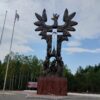 Bronze memorial sculpture resembling a tree with winged figures and inscribed names, part of the Wołyń massacre monument, with flagpoles nearby.