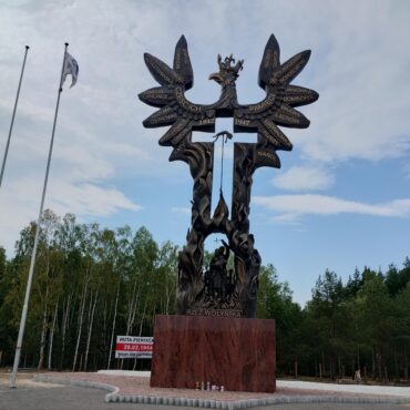 Bronze memorial sculpture resembling a tree with winged figures and inscribed names, part of the Wołyń massacre monument, with flagpoles nearby.