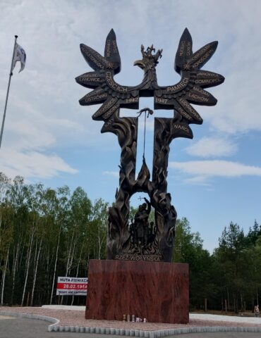 Bronze memorial sculpture resembling a tree with winged figures and inscribed names, part of the Wołyń massacre monument, with flagpoles nearby.