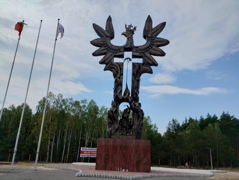 Bronze memorial sculpture resembling a tree with winged figures and inscribed names, part of the Wołyń massacre monument, with flagpoles nearby.