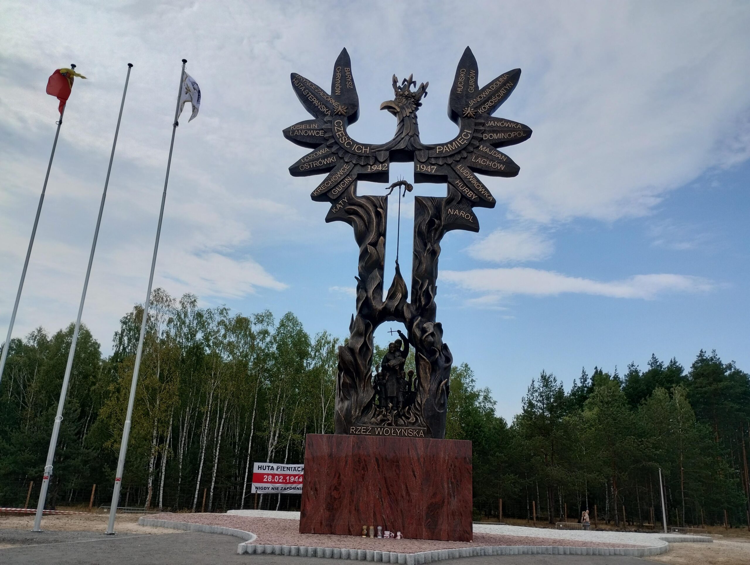 Bronze memorial sculpture resembling a tree with winged figures and inscribed names, part of the Wołyń massacre monument, with flagpoles nearby.
