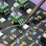 A soldering iron touching a circuit board pad near small green tactile switches and white LEDs on a blue/black PCB filled with labeled traces and golden pad holes.