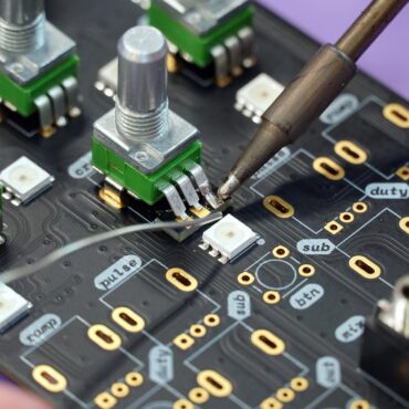 A soldering iron touching a circuit board pad near small green tactile switches and white LEDs on a blue/black PCB filled with labeled traces and golden pad holes.