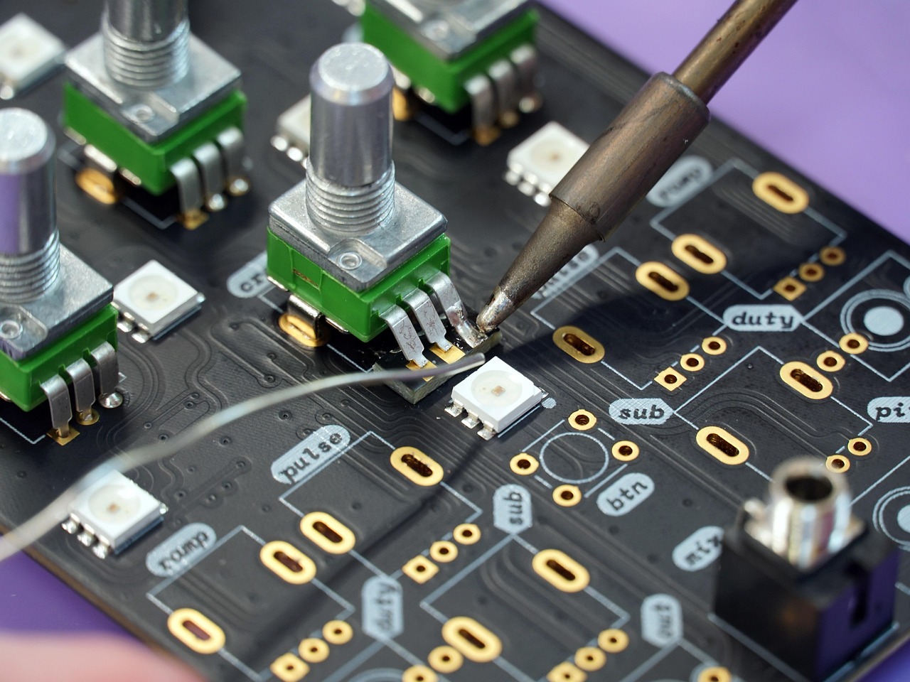 A soldering iron touching a circuit board pad near small green tactile switches and white LEDs on a blue/black PCB filled with labeled traces and golden pad holes.