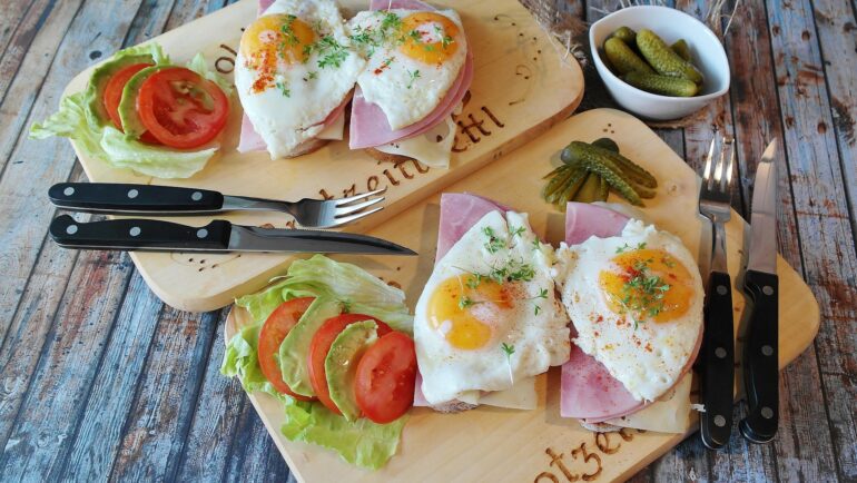 jajka - NOTE.radio - Polskie radio w UK | Polskie radio w Londynie Two open-face breakfast sandwiches with ham, cheese and sunny-side-up eggs, garnished with herbs, on a wooden board with tomato, lettuce and pickles nearby.