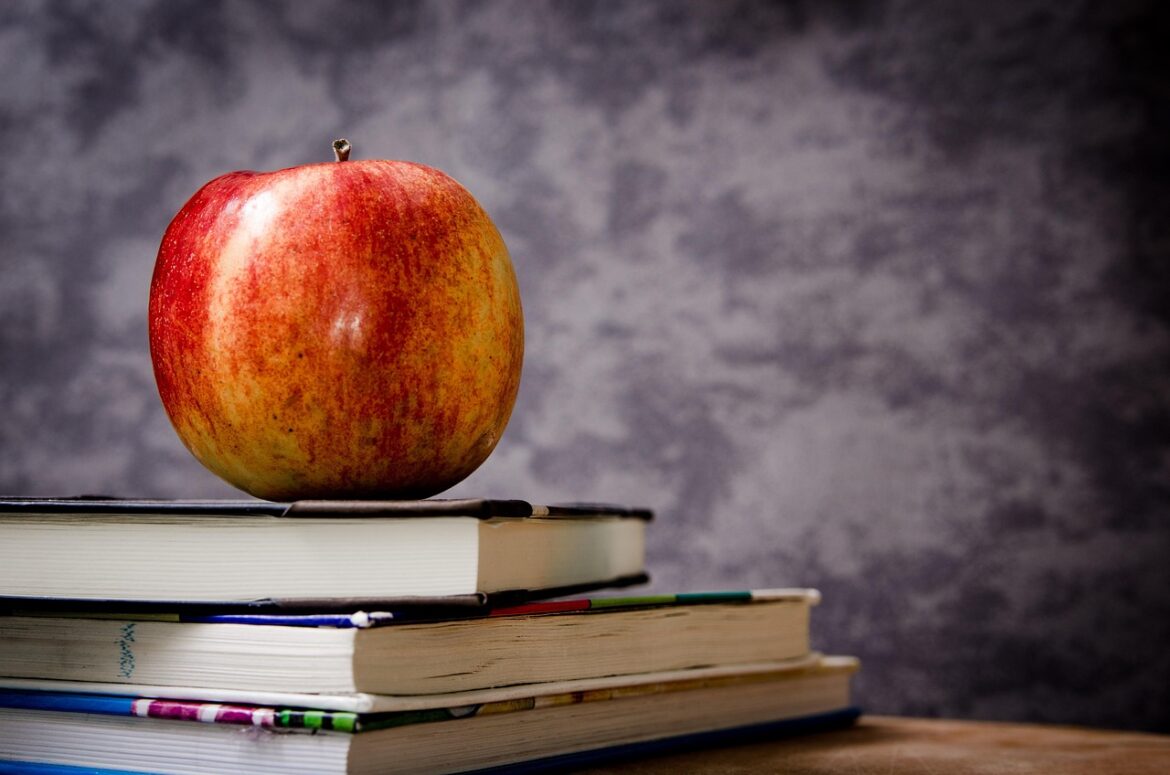 Red and yellow apple resting on top of a stack of closed books on a wooden table.