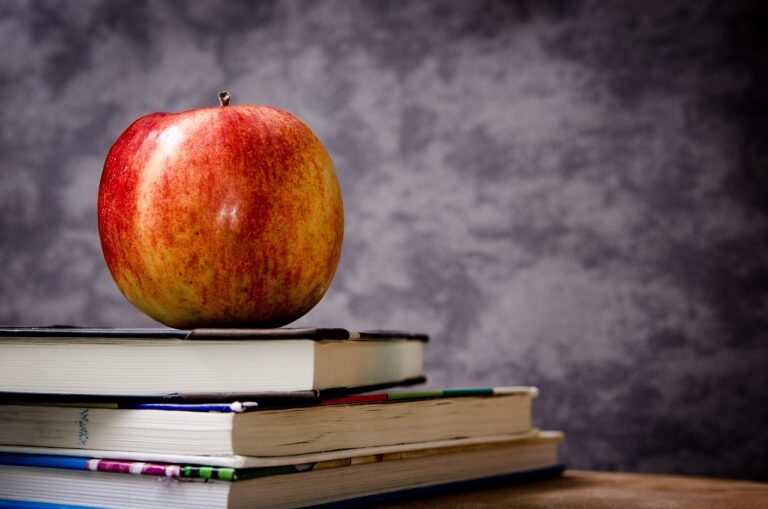 Red and yellow apple resting on top of a stack of closed books on a wooden table.