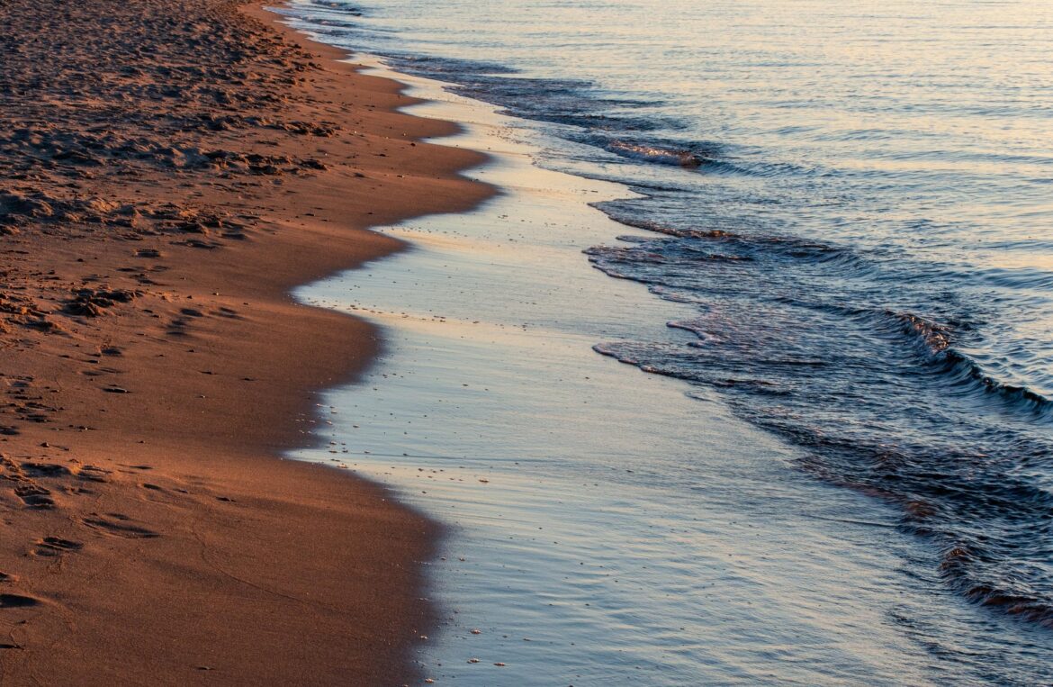 Sunlit sandy beach meeting calm ocean waves, with a gentle shoreline curving into the distance at sunset.