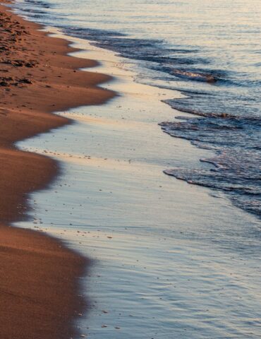 Sunlit sandy beach meeting calm ocean waves, with a gentle shoreline curving into the distance at sunset.