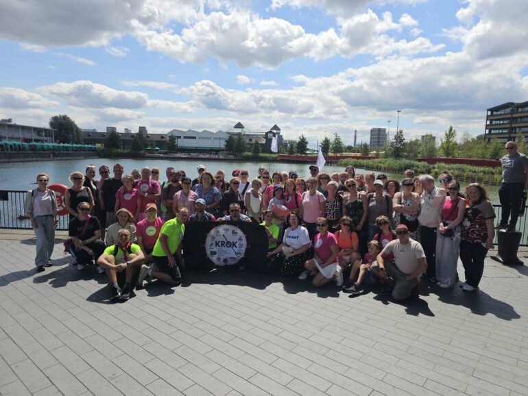 1000071885 - NOTE.radio - Polskie radio w UK | Polskie radio w Londynie Large group of people posing together by a waterfront with modern buildings and a blue sky with clouds in the background. many wearing bright pink and neon shirts, some with badges, forming a crowd on a paved promenade over the water.