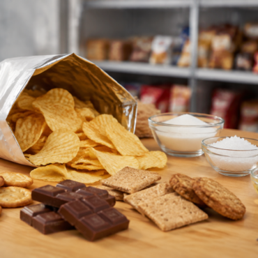 Open bag of potato chips spilling onto a wooden table with crackers, cookies, chocolate, and bowls of sugar and flour nearby.